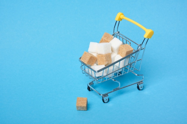 Cubetti di zucchero bianco e marrone in un carrello della spesa su uno spazio di copia superficie blu