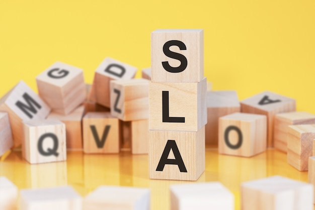 Cubi di legno con lettere SLA disposti in una piramide verticale, sfondo giallo, riflesso dalla superficie del tavolo, concetto di business, SLA - abbreviazione di accordo sul livello di servizio