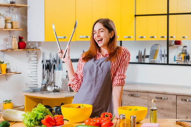 Cucina casalinga divertente Signora allegra in piedi con le pinze da cucina sorridente che strizza l'occhio mentre prepara insalata di verdure