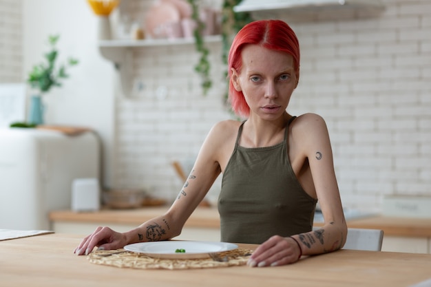 Cucina da solo. Donna anoressica dai capelli rossi che ha un aspetto orribile mentre è seduta in cucina da sola