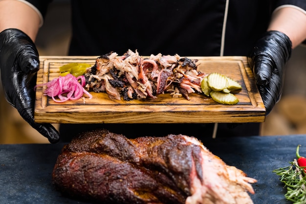 Cucina del ristorante grill. Mani dello chef in guanti da cucina neri che tengono carne di maiale affumicata con sottaceti su tavola di legno.