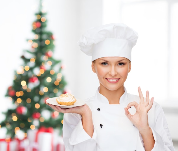 Cucina, persone, vacanze e concetto di cibo - chef, cuoca o fornaio sorridente con cupcake sul piatto che mostra un gesto ok sul soggiorno e sullo sfondo dell'albero di natale