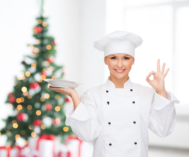 cucina, persone, vacanze e concetto di cibo - sorridente chef, cuoco o fornaio con piatto vuoto che mostra ok segno sul soggiorno e sullo sfondo dell'albero di natale