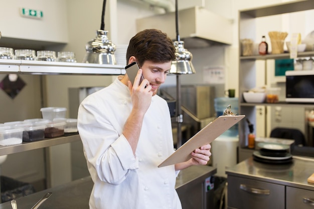 cucina, professione e concetto di persone - cuoco cuoco con clipboard che chiama sullo smartphone in cucina del ristorante