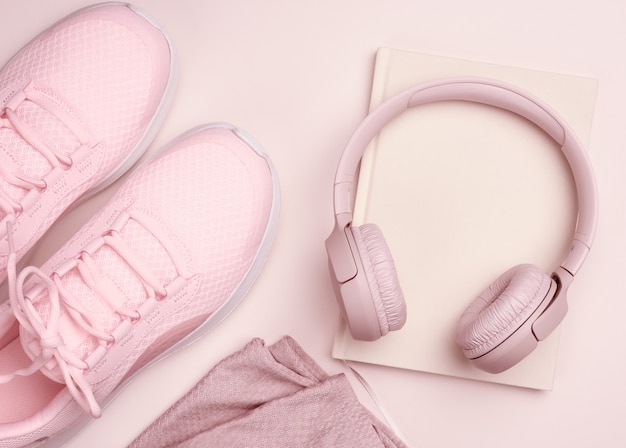 Cuffie wireless rosa, un paio di scarpe da ginnastica e un blocco note su fondo beige, vista dall'alto. Abbigliamento Donna