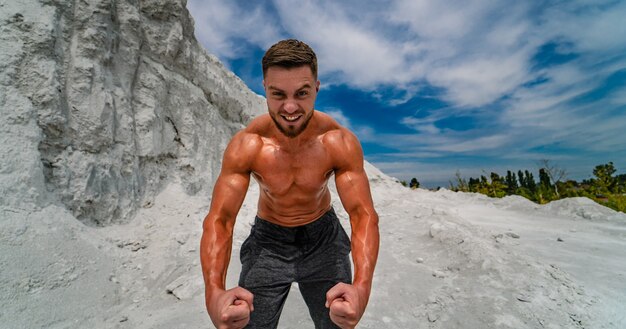 Culturista con perfetto, spalle, bicipiti, tricipiti e petto in posa all'aperto sullo sfondo del cielo blu. Uomo forte e atletico in piedi con il torso nudo. Concetto di bel corpo sano.