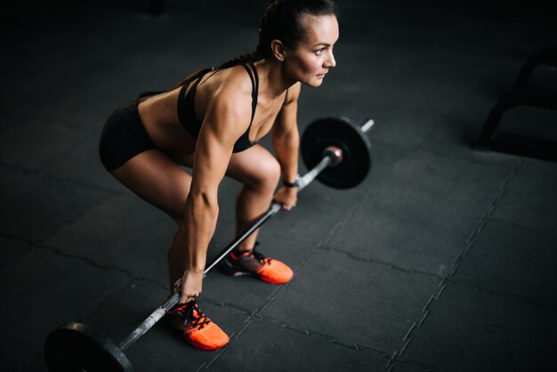 Culturista donna forte con un corpo fitness perfetto che si prepara a sollevare il pesante bilanciere dal pavimento. Concetto di stile di vita sano e allenamenti in una moderna palestra buia.