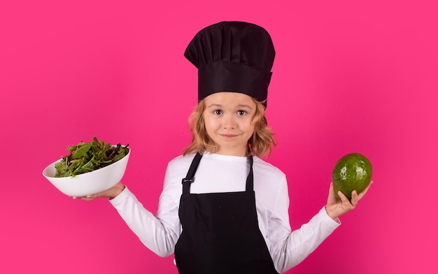 Cuoco bambino divertente con avocado e verdura Bambino che indossa uniforme da cucina e cappello da chef che prepara il cibo in cucina ritratto in studio Cucinare il concetto di cucina e cibo per bambini