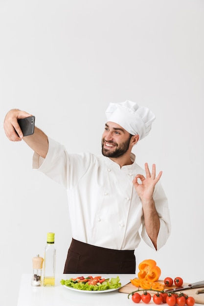 Cuoco caucasico uomo in uniforme che mostra segno ok e scatta selfie foto di cibo sullo smartphone al lavoro isolato su muro bianco