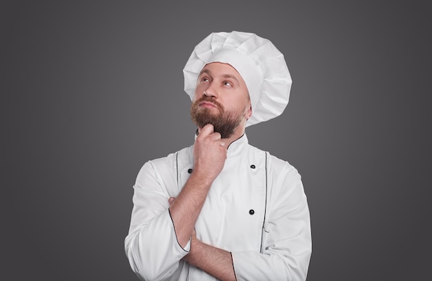 Cuoco maschio adulto premuroso barbuto in uniforme bianca da chef e cappello strofinando il mento e guardando in alto mentre si pensa al menu, nella cucina del ristorante su sfondo grigio