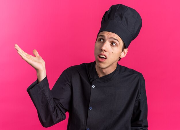 Cuoco maschio giovane biondo senza indizi in uniforme da chef e berretto che guarda in alto mostrando la mano vuota isolata sulla parete rosa