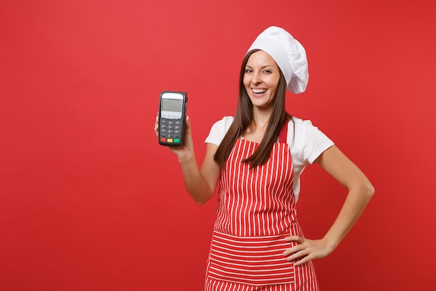 Cuoco o fornaio femminile della casalinga in cappello da cuoco unico del toque della maglietta del grembiule a strisce isolato sul fondo rosso della parete. La donna tiene in mano il dispositivo nfc del terminale di pagamento bancario wireless. Deridere il concetto dello spazio della copia.