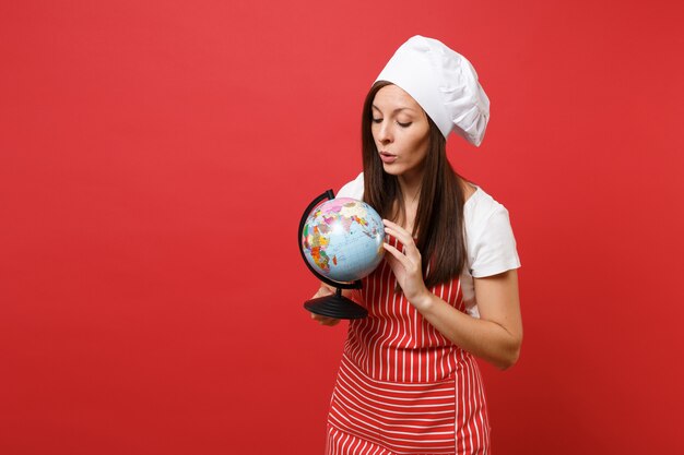 Cuoco o fornaio femminile della casalinga in grembiule a strisce, maglietta bianca, cappello del cuoco unico di toque isolato sul fondo rosso della parete. Donna della governante che tiene nel globo del mondo della terra delle palme. Deridere il concetto dello spazio della copia.