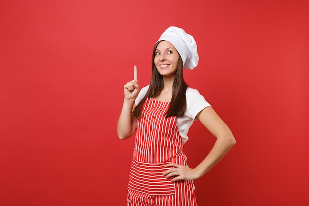 Cuoco o fornaio femminile della casalinga in grembiule a strisce, t-shirt bianca, cappello da cuoco unico del toque isolato sul fondo rosso della parete. Bella donna della governante che punta il dito indice verso l'alto. Deridere il concetto dello spazio della copia.