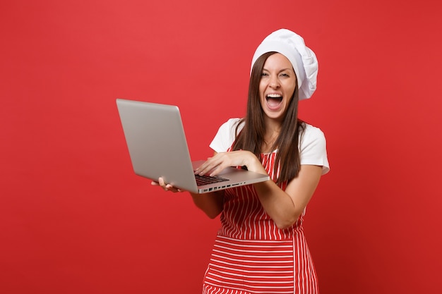 Cuoco o fornaio femminile della casalinga in grembiule a strisce, t-shirt bianca, cappello da cuoco unico del toque isolato sul fondo rosso della parete. Donna governante divertente che cerca la ricetta nel pc portatile. Deridere il concetto dello spazio della copia.