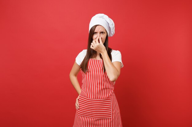 Cuoco o fornaio femminile della casalinga in grembiule a strisce, t-shirt bianca, cappello da cuoco unico del toque isolato sul fondo rosso della parete. La donna della governante chiude il naso con la mano perché puzza. Deridere il concetto dello spazio della copia.