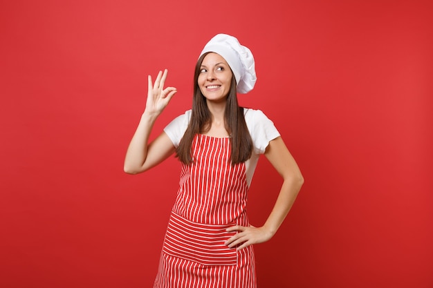 Cuoco o fornaio femminile della casalinga in maglietta bianca del grembiule a strisce, cappello del cuoco unico di toque isolato sul fondo rosso della parete. Sorridente donna carina calma che fa segno di delizia di gusto ok. Deridere il concetto dello spazio della copia.