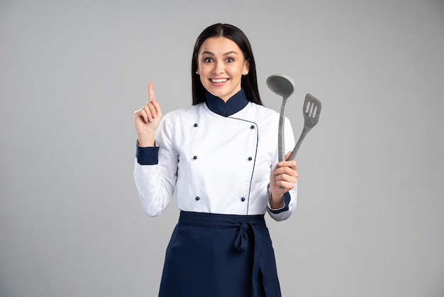 Cuoco o fornaio femminile della casalinga in uniforme che tiene dito su e che ha idea isolata sul fondo grigio della parete. Foto d'archivio