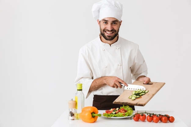 cuoco professionista uomo in uniforme sorridente e taglio insalata di verdure su tavola di legno isolato su muro bianco