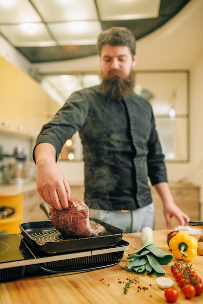 Cuoco unico barbuto che cucina la carne in una padella sulla cucina. Uomo che prepara carne di maiale bollita sul fornello elettrico da tavola