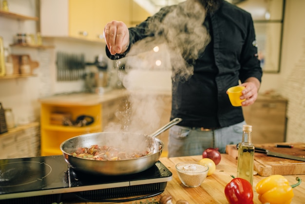 Cuoco unico maschio salato carne con vetables nella padella in cucina. Uomo che prepara carne bovina sul fornello elettrico da tavola