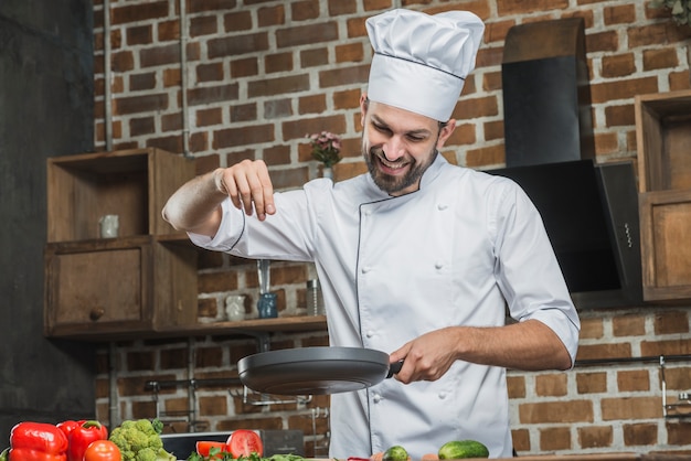 Cuoco unico sorridente che sta nella cucina che spruzza le spezie sulla padella