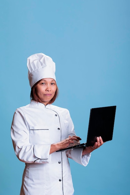 Cuoco unico sorridente con il grembiule che tiene il computer portatile che controlla la lista degli ingredienti della ricetta culinaria, preparando il piatto di gastronomia. Persona asiatica allegra che lavora nell'industria alimentare che cucina un pasto sano