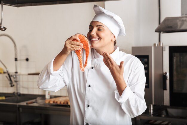 Cuoco unico sorridente della donna che indossa l'uniforme che cucina la bistecca di color salmone delizioso che sta alla cucina