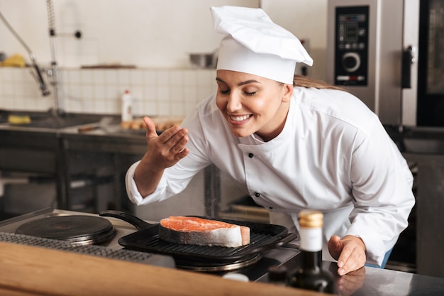 Cuoco unico sorridente della donna che indossa l'uniforme che cucina la bistecca di color salmone delizioso che sta alla cucina