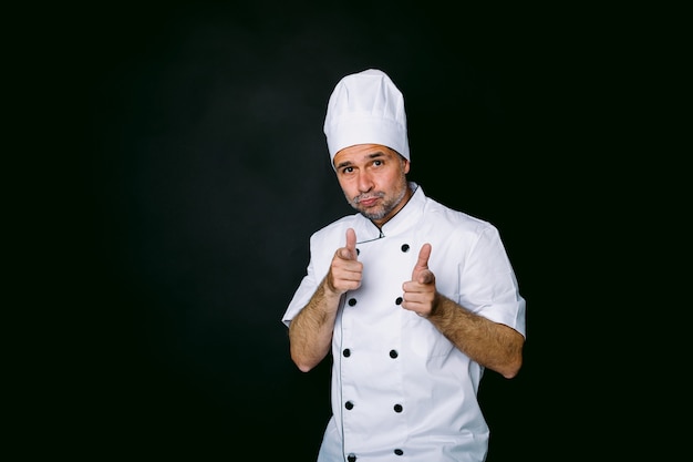 Cuoco vestito con giacca e cappello bianco alzando le mani in segno di approvazione, su sfondo nero. Ristorante e concetto da asporto.