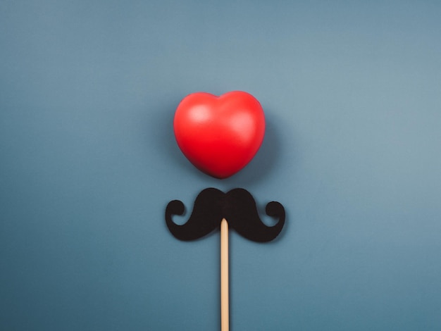 Cuore rosso palla e baffi bastone isolato su sfondo blu, stile minimal. Buona festa del papà, concetto Movember.