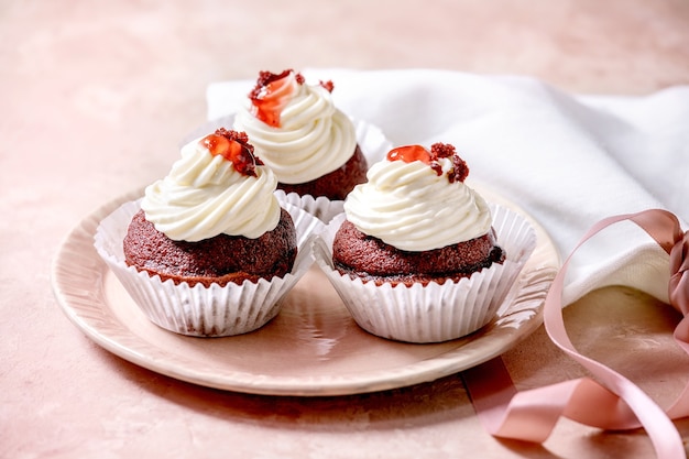 Cupcakes di velluto rosso fatti in casa con panna montata sul piatto in ceramica rosa, tovagliolo bianco con nastro sopra la parete di struttura rosa