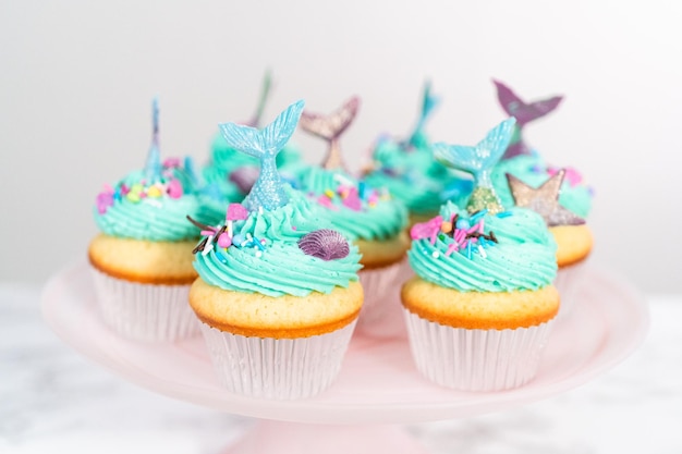 Cupcakes gourmet a forma di sirena conditi con glassa di crema al burro blu e decorati con confettini e code di sirena al cioccolato.