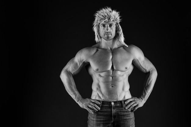 Cura del corpo con fitness e sport Uomo con torso muscoloso Bodybuilder in cappello di pelliccia e jeans sfondo scuro Cura del corpo e benessere Salute e dieta