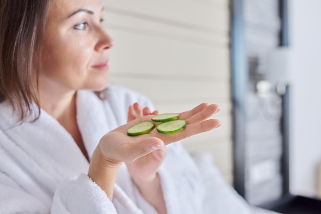 Cura della pelle del viso di una donna di mezza età. Donna matura in accappatoio che fa trattamenti termali domestici con cetrioli, cosmetici biologici naturali e massaggio facciale antietà a casa a letto