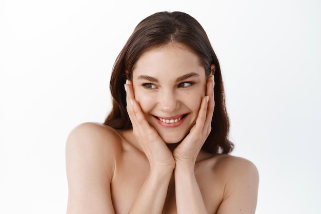 Cura della pelle e bellezza Ragazza con pelle nutrita e pulita che tocca il suo viso pulito e idratato sorridente e distoglie lo sguardo Donna con spalle nude utilizzando cosmetici per la cura della pelle sfondo bianco