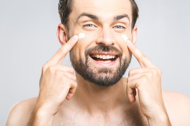 Cura della pelle. Giovane uomo senza camicia bello che applica crema al suo fronte e che si esamina mentre controllando fondo grigio