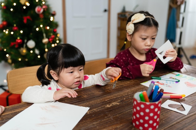 curiosa bambina asiatica impegnata nella creatività con sua sorella per le vacanze di Natale sta guardando le forbici in mano con curiosità mentre impara a usarle