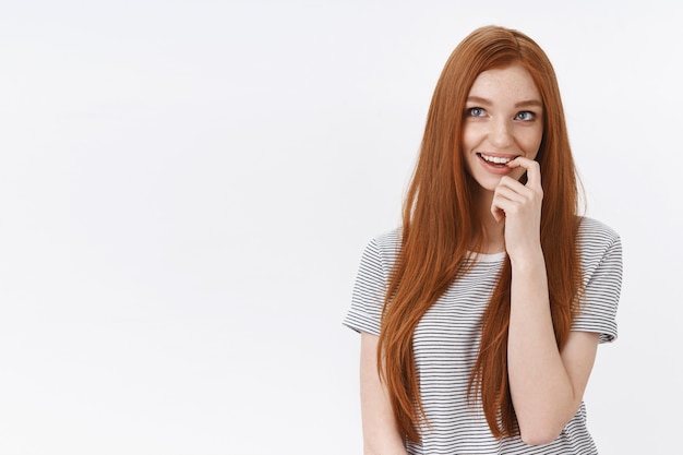 Curioso incuriosito adorabile giovane ragazza rossa romantica occhi azzurri che morde il dito sorridente felice immagina un'idea interessante pensando guarda nell'angolo in alto a sinistra avere desiderio, muro bianco