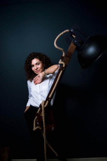 Curly giovane donna in abiti da lavoro in posa da lampada in legno elemento interno del dispositivo