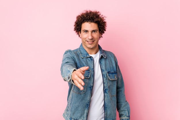 Curly uomo maturo che indossa una giacca di jeans contro il muro rosa che allunga la mano alla macchina fotografica nel gesto di saluto.