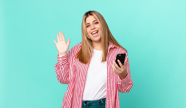 Curvy bella donna bionda che sorride felicemente, agitando la mano, accogliendoti e salutandoti e tenendo in mano uno smartphone