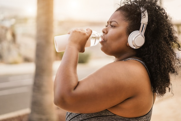 Curvy donna nera che beve acqua dopo la routine di jogging all'aperto nel parco cittadino - Focus on face