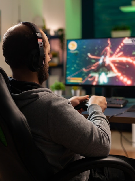 Cyber man con auricolare che gioca in competizione con controller wireless in uno studio professionale attrezzato. Giocatore entusiasta seduto sulla sedia da gioco guardando il monitor per vincere il campionato online