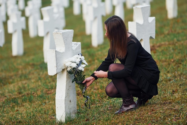 Dà rispetto mettendo fiori Giovane donna in abiti neri in visita al cimitero con molte croci bianche Concezione del funerale e della morte