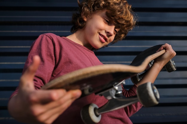 Da sotto un piccolo pattinatore positivo che sorride e controlla lo skateboard mentre si trova vicino al muro blu scuro in città