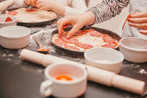 Da vicino i bambini stendono pezzi di salame sulla base della pizza