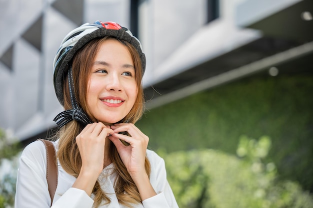 Da vicino una donna sorridente che indossa un casco che si esercita all'aperto con la bicicletta in strada una giovane donna d'affari asiatica che mette il casco da bicicletta per la sicurezza preparati ciclisti intorno all'edificio vanno al lavoro in bicicletta