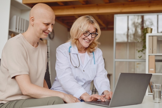 Dai toni caldi ritratto di allegro medico donna utilizzando laptop e parlando con paziente calvo durante la consultazione su alopecia e recupero del cancro, spazio di copia