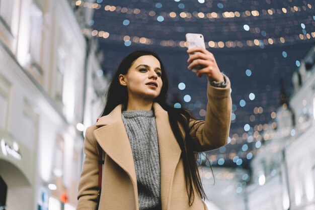 Dal basso di una giovane donna alla moda in cappotto in piedi e che si fa selfie con lo smartphone